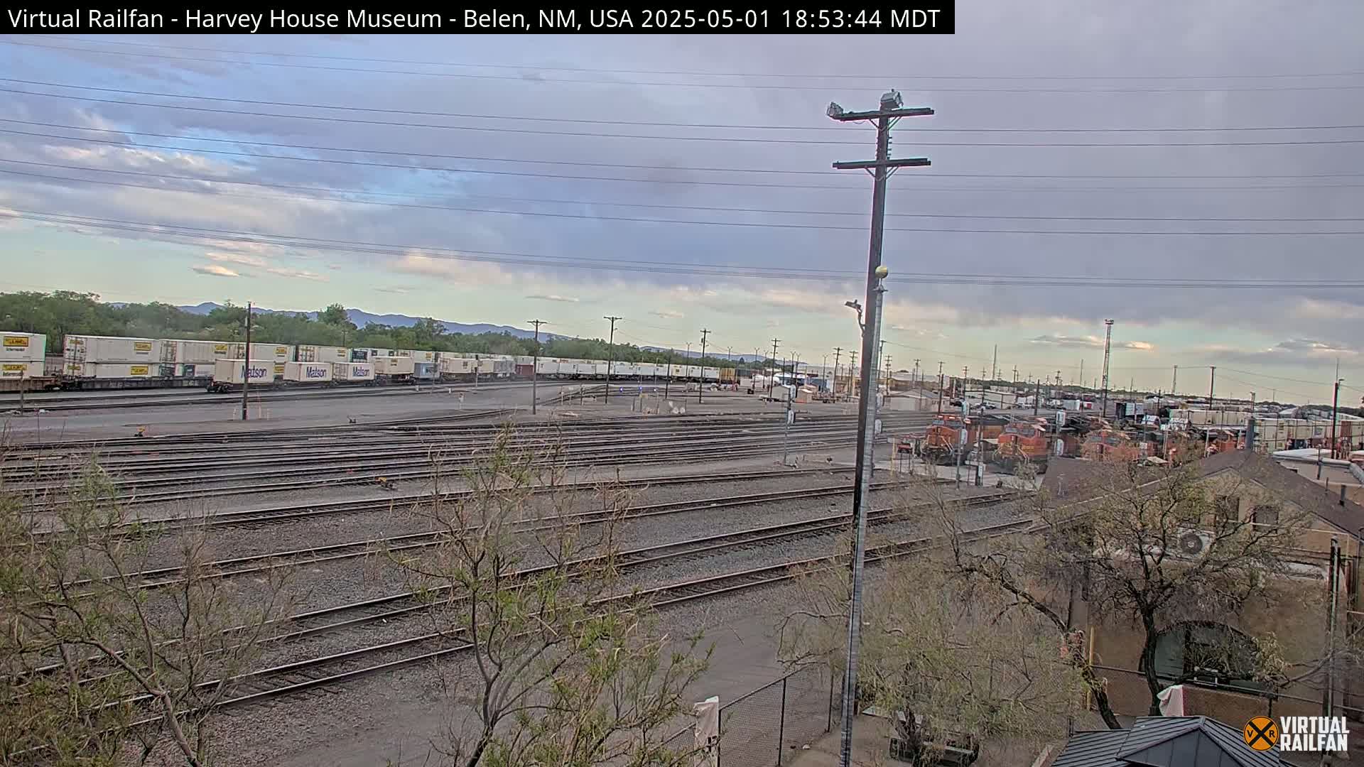 A sprawling rail yard with numerous train tracks, a collection of boxcars, and several locomotives under a partly cloudy sky.