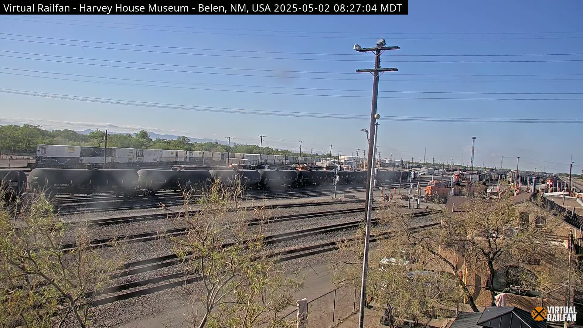 Under a clear blue sky, a long line of black train cars sits on a railway track, with other trains and industrial buildings visible in the background.