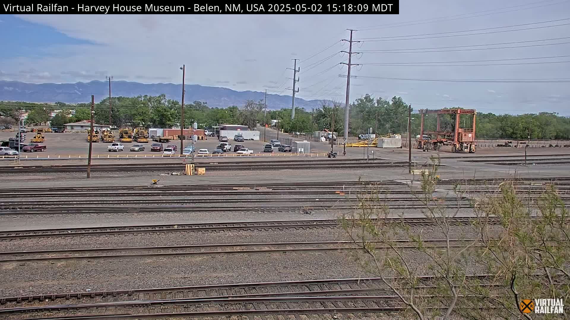 A railway yard with numerous train tracks, parked vehicles, and construction equipment, set against a backdrop of mountains under a partly cloudy sky.