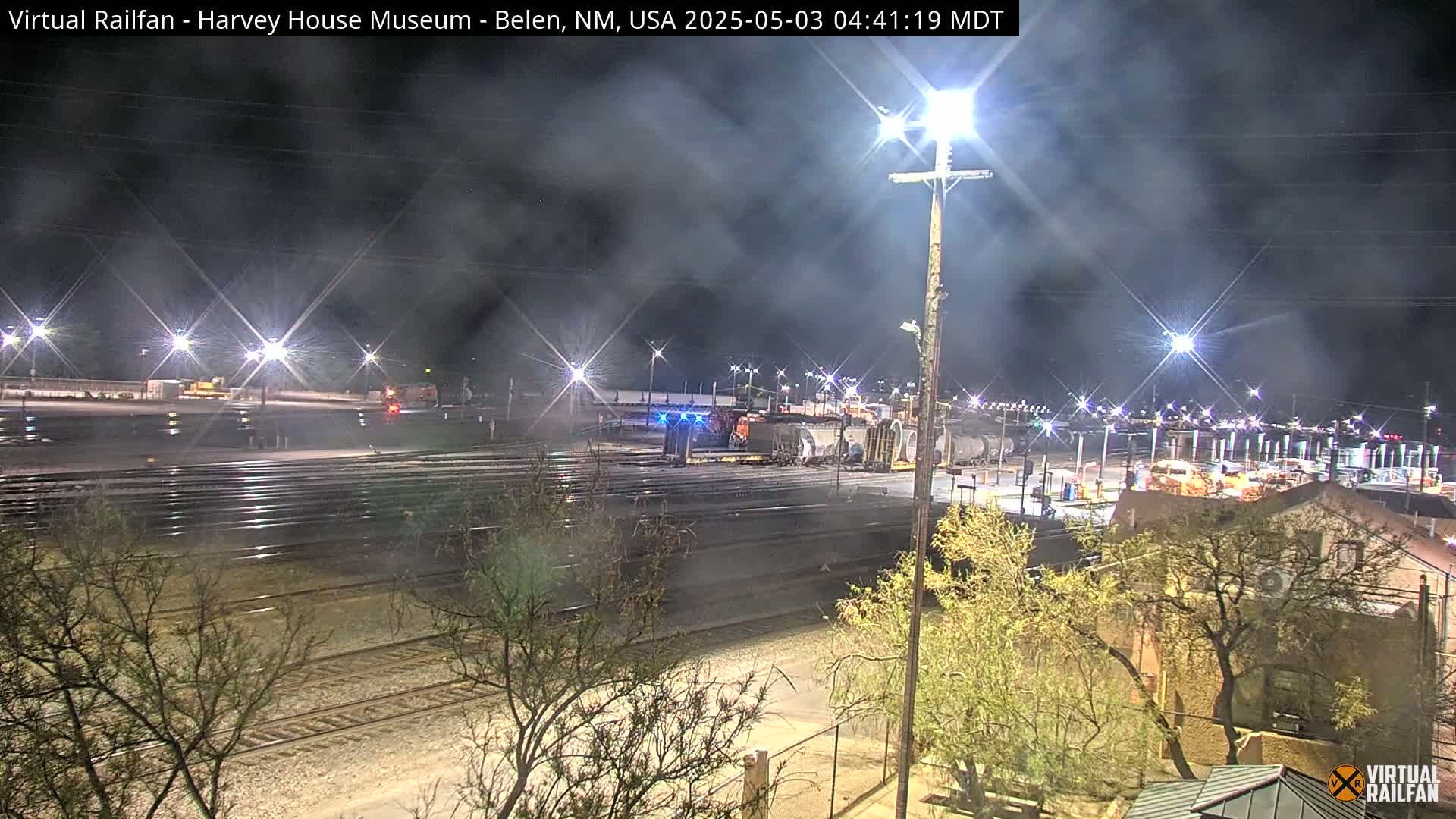 A nighttime view of a train yard with several rail lines, some train cars, and surrounding buildings under hazy conditions.