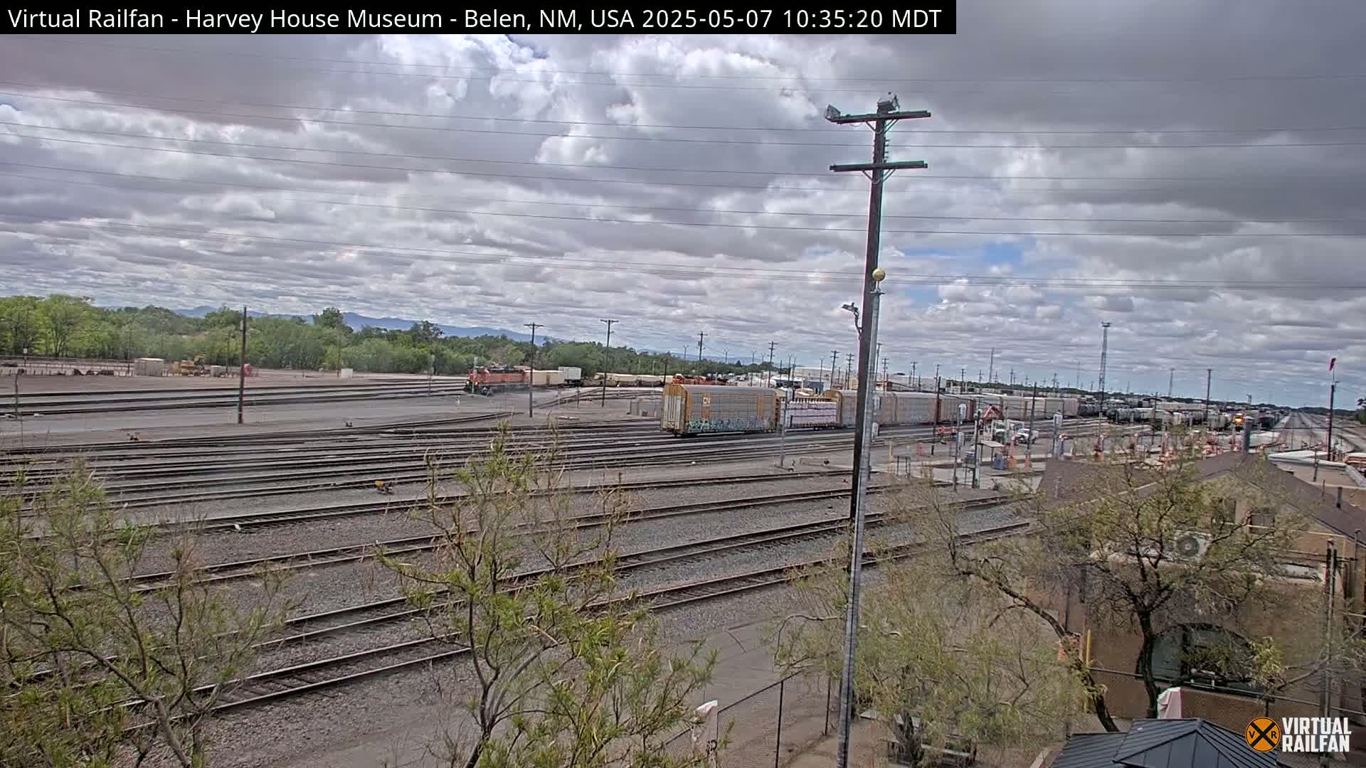 A wide shot of a railway yard with many train cars and tracks under a partly cloudy sky.