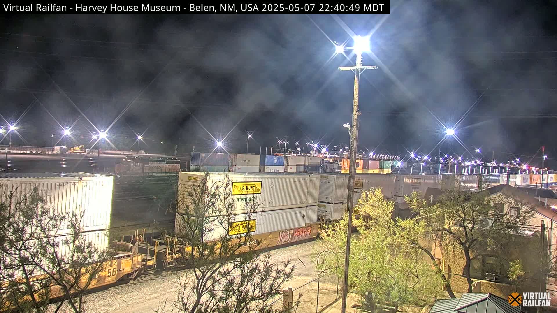 A nighttime view of a train carrying numerous shipping containers in a rail yard, under a cloudy sky illuminated by bright lights.