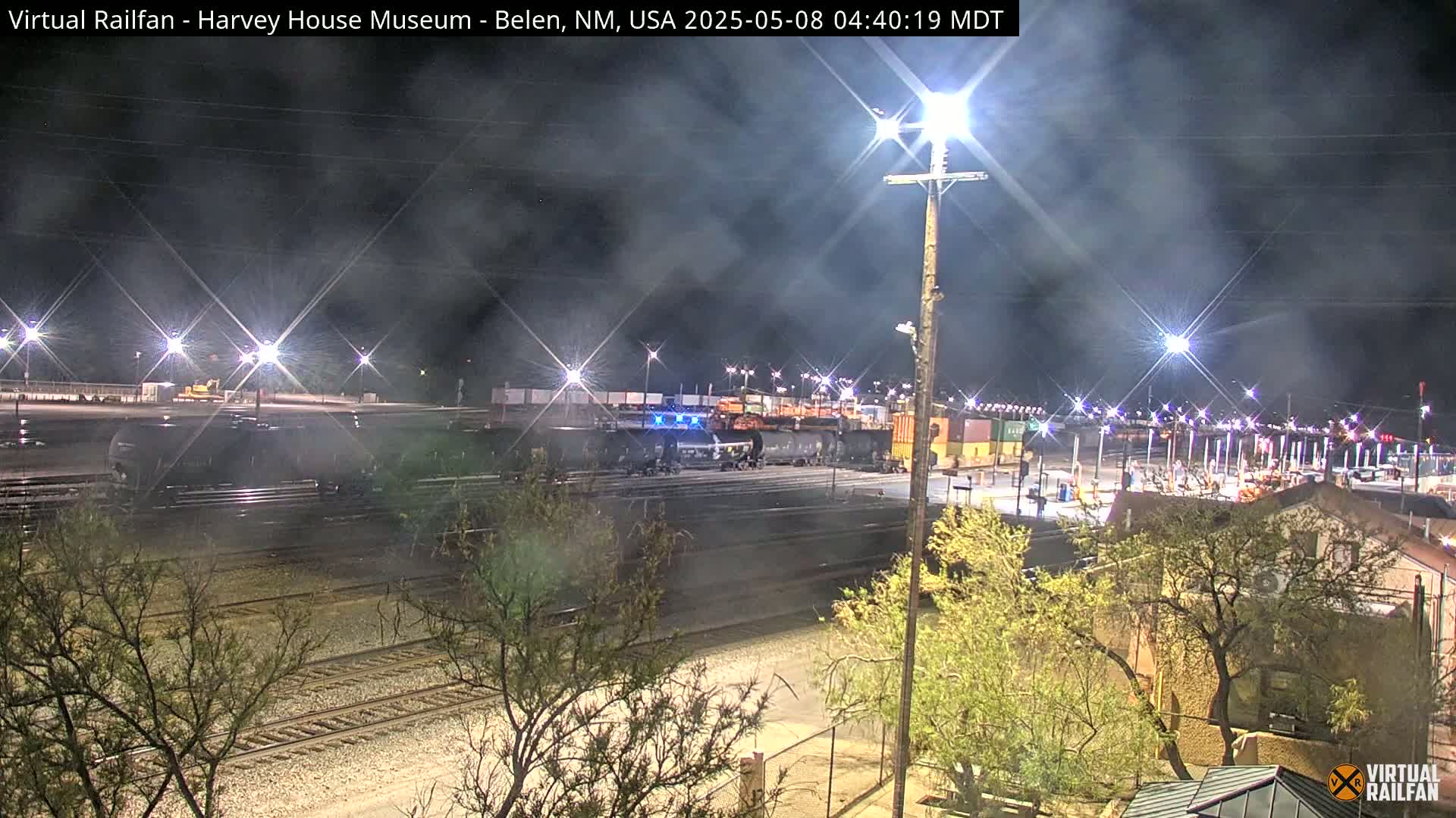 A nighttime view of a train yard with several rail cars and a locomotive, under a hazy sky illuminated by bright lights.