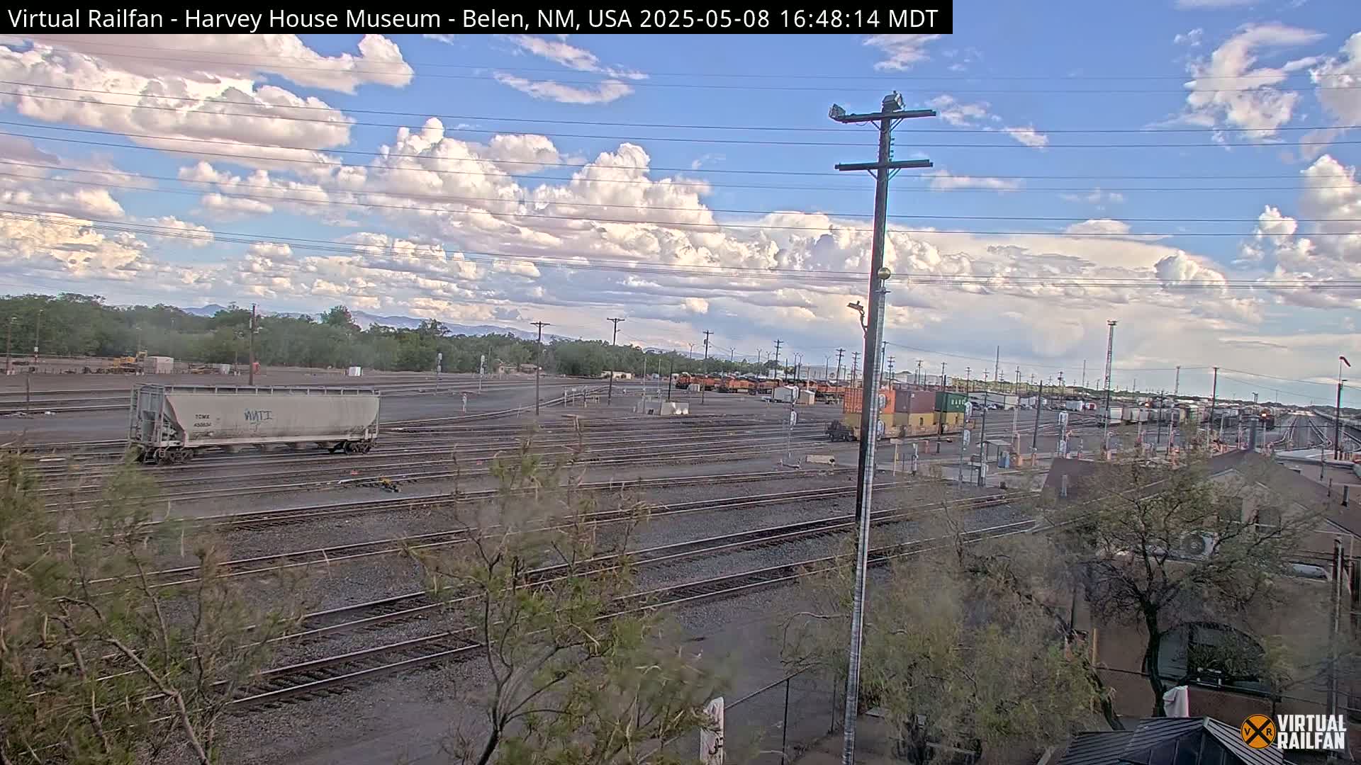 A partly cloudy sky overlooks a large rail yard with multiple train cars and tracks under a mostly sunny sky.