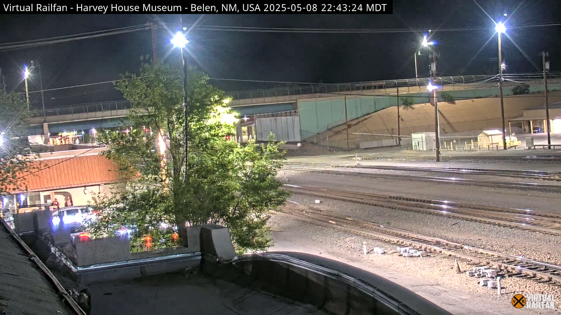 A nighttime view of railroad tracks and a roadway under a bridge, with some cars and buildings visible.