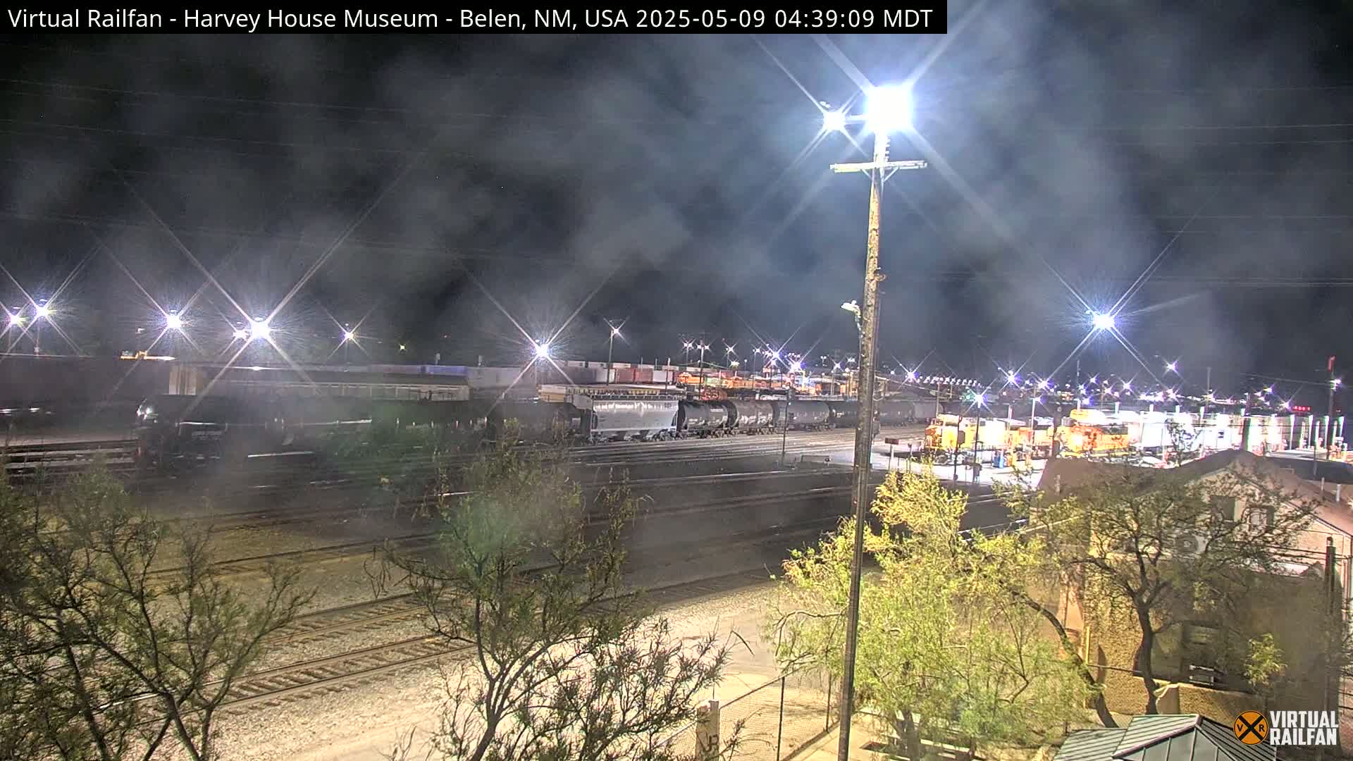 A nighttime view of a train yard with numerous railcars and locomotives under a hazy, dark sky.