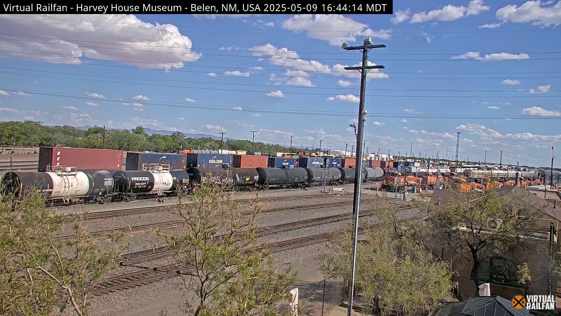 A long train of tanker cars and shipping containers sits on a railway track under a partly cloudy sky.