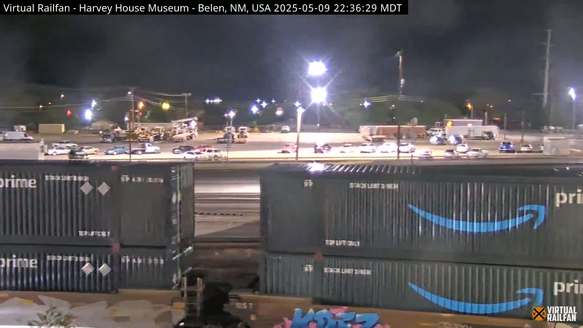 A nighttime view of a train carrying Amazon Prime shipping containers, parked next to a large, brightly lit parking lot with numerous vehicles.