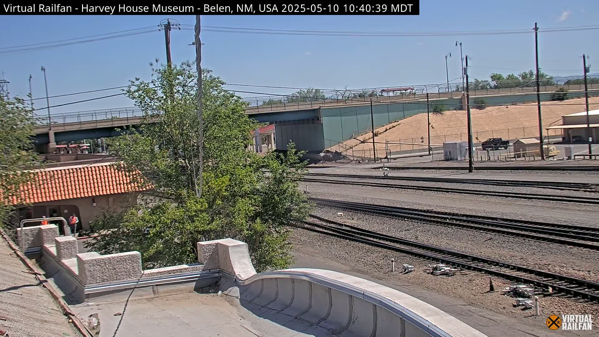 A view of railroad tracks and a road overpass under a clear, sunny sky, with a building and some trees in the foreground.
