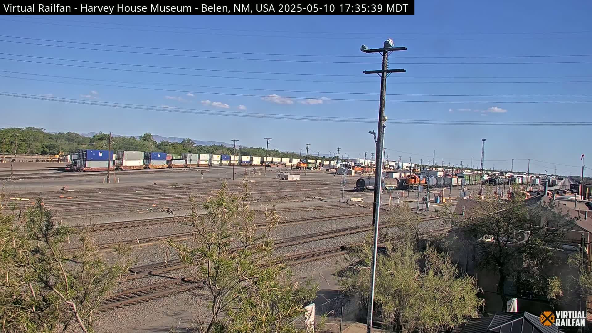 Under a clear blue sky, a large rail yard with numerous train cars and containers is shown, alongside a few small buildings and sparse vegetation.
