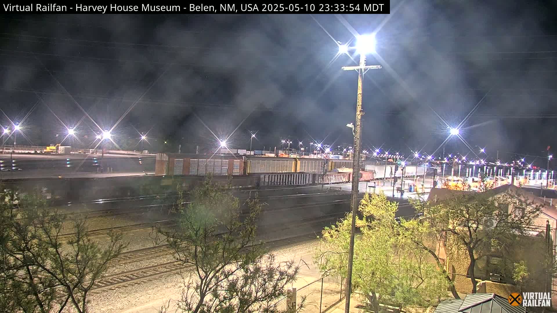A nighttime view of a train yard with a long freight train, illuminated by bright lights, under a hazy, dark sky.