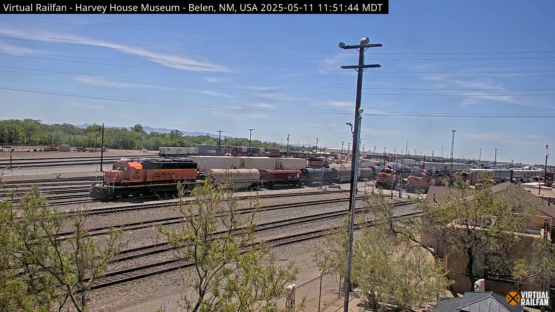 An orange locomotive leads a train of freight cars in a sunny, outdoor rail yard.