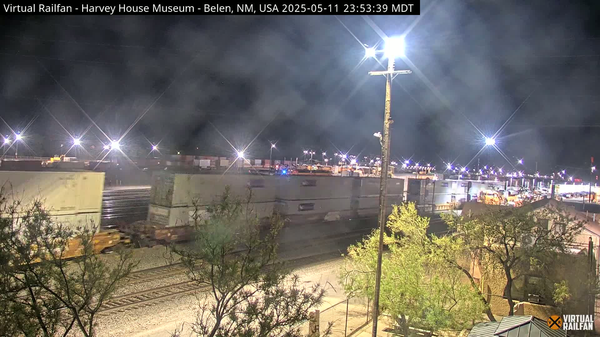 A nighttime view of a train yard with freight cars and containers, under a slightly hazy sky illuminated by bright lights.
