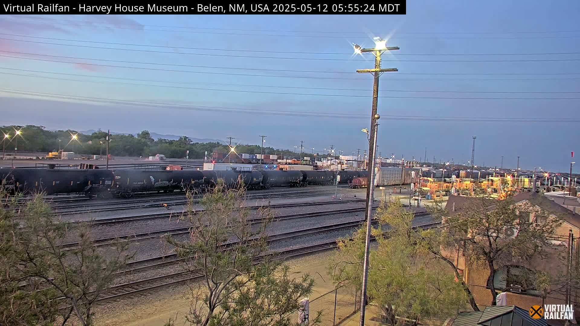 Under a twilight sky, several dark freight cars sit on a network of train tracks near a building and some trees.