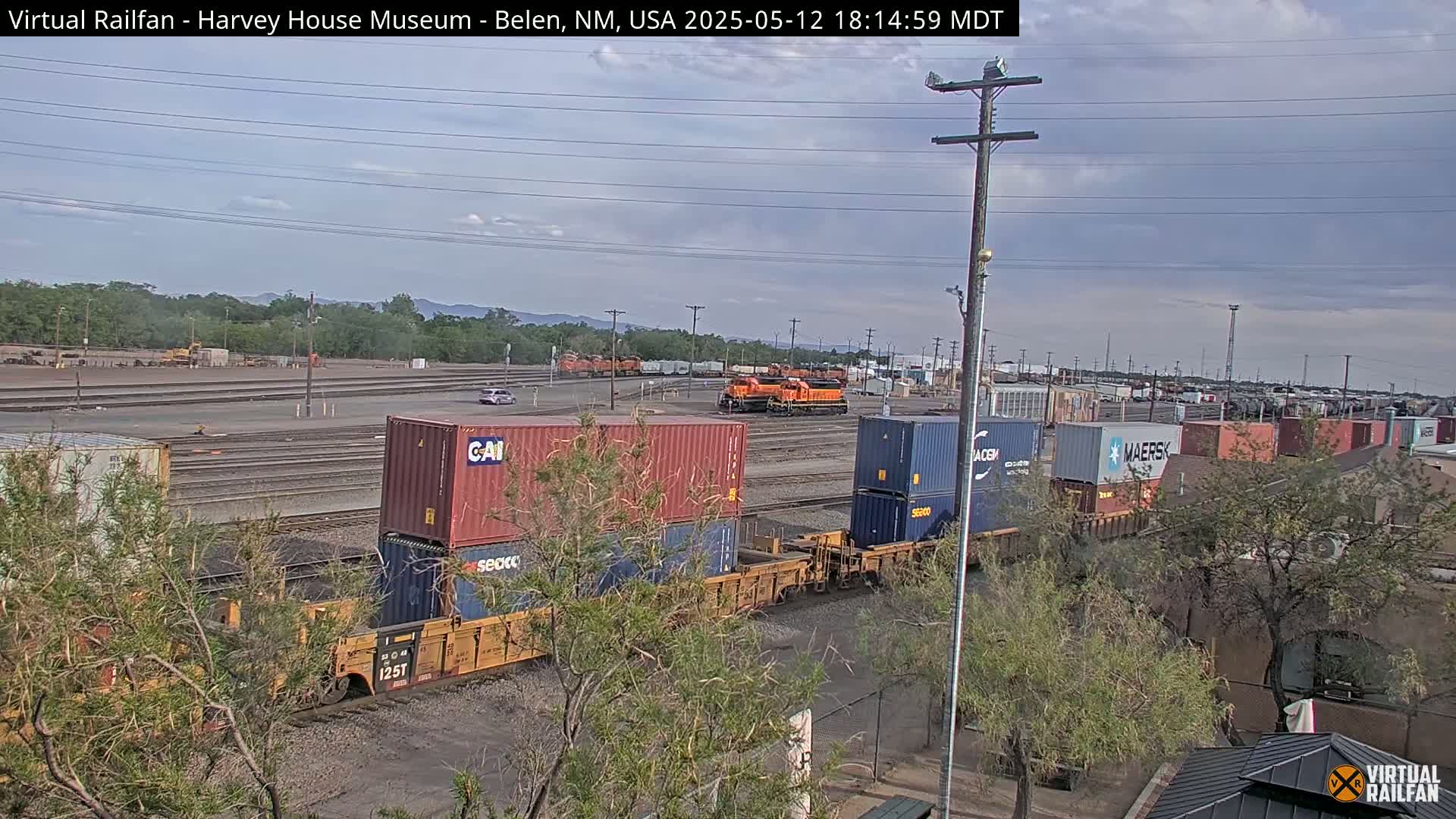 A mostly clear sky above a train yard with an orange locomotive and several shipping containers on railcars.