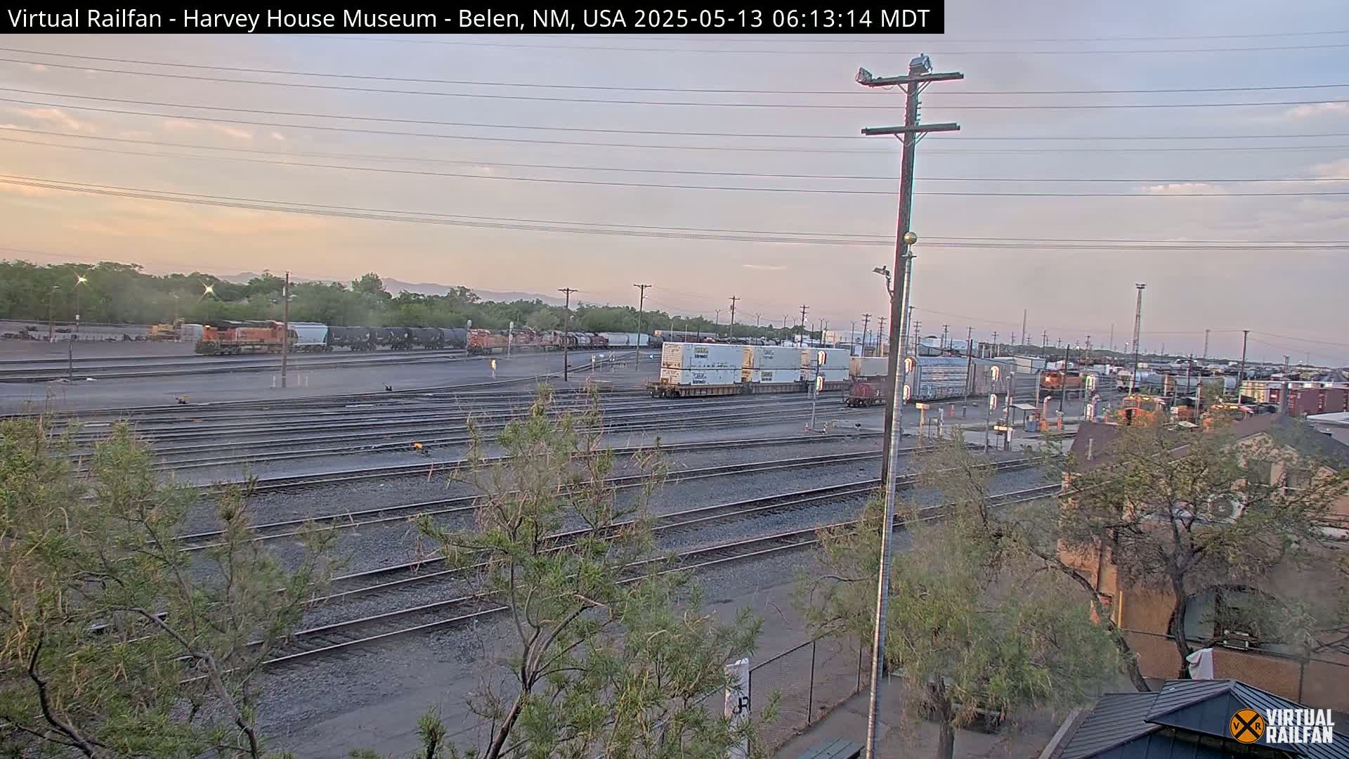 Under a soft, pale sunrise, a sprawling railroad yard displays numerous train cars and tracks, with several locomotives visible among them.