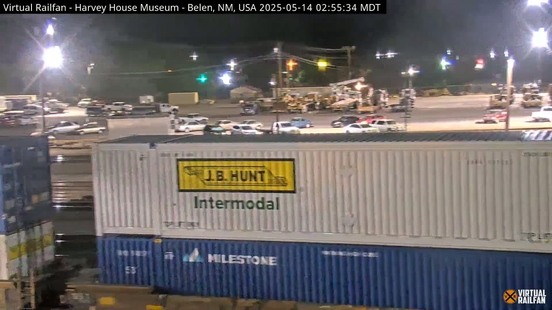 At night, under bright lights, a train car with shipping containers sits in a parking lot surrounded by other cars.