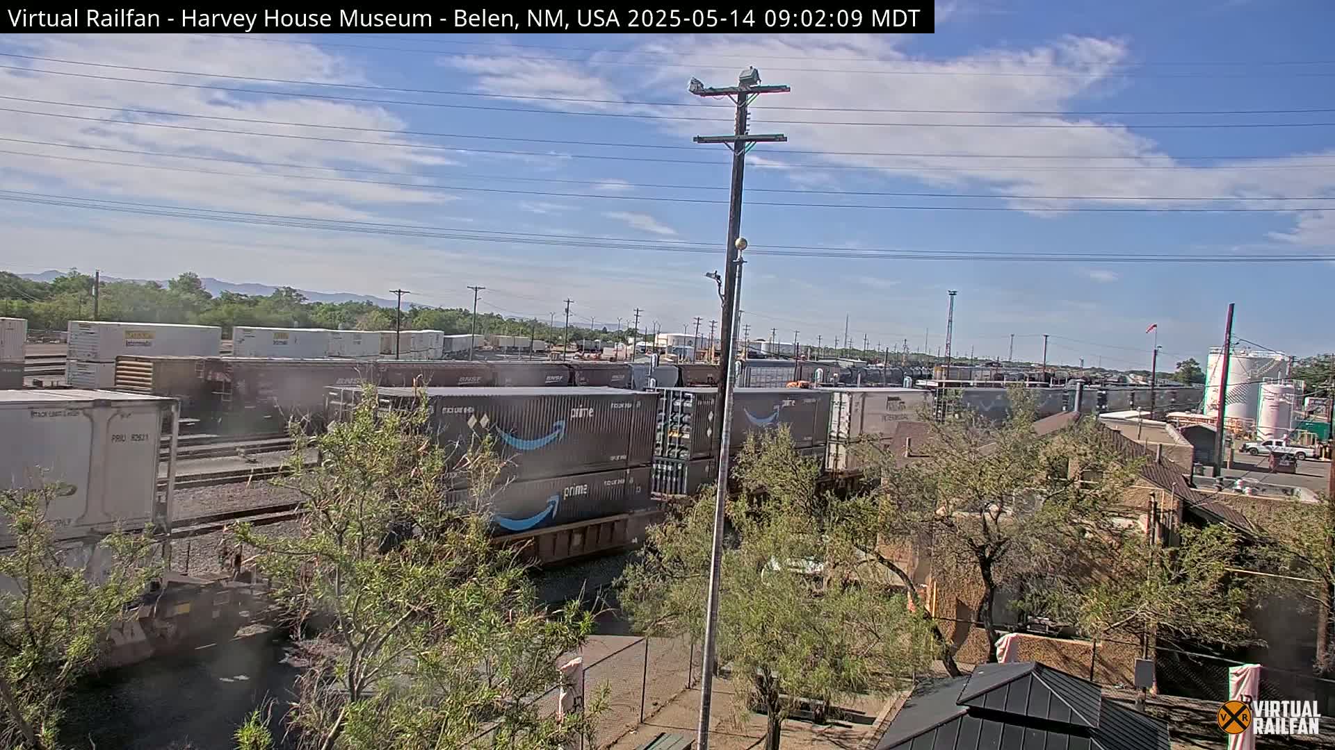 A partly cloudy day reveals a freight train carrying Amazon containers, situated near a building and trees, under a power line.