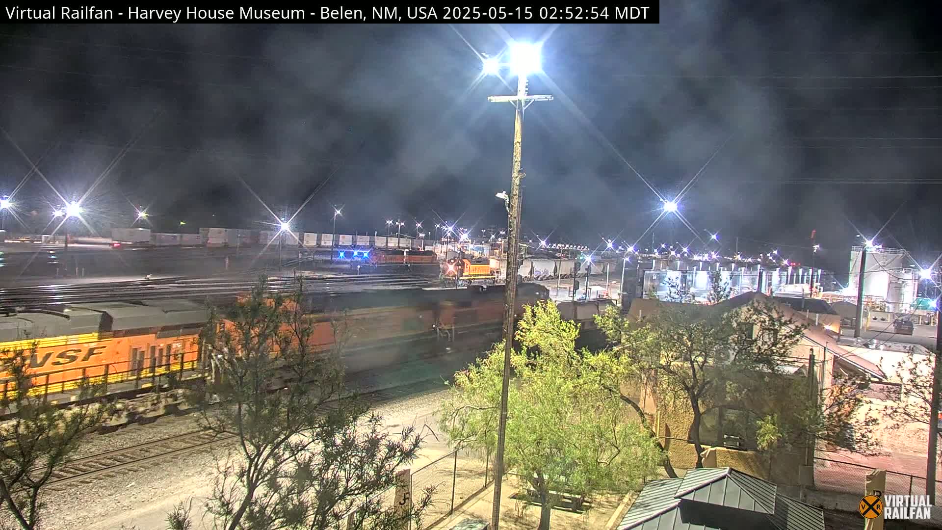 A long freight train passes through a brightly lit rail yard at night under a mostly clear sky.
