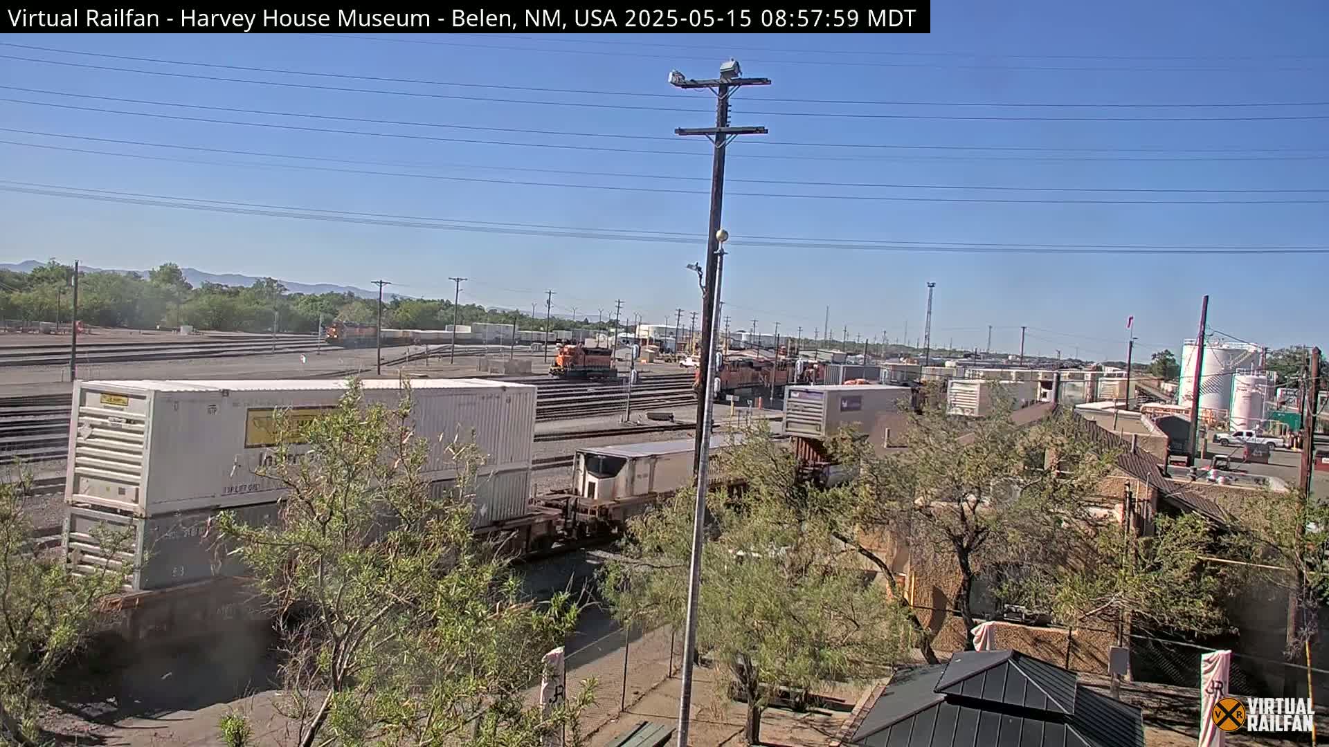 A sunny day reveals a train yard with multiple train cars and containers, partially obscured by trees in the foreground.