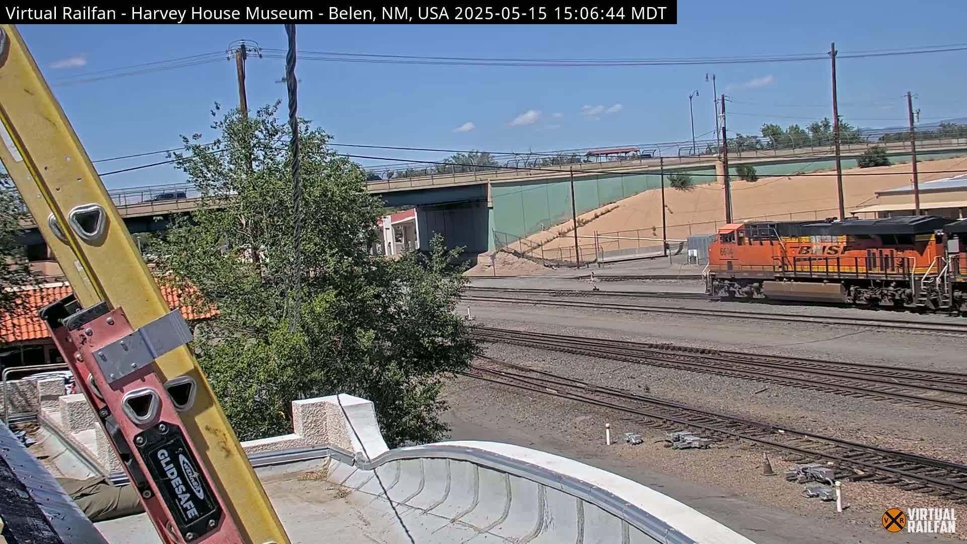 An orange locomotive is seen traveling on train tracks under a sunny sky, with a ladder and part of a building visible in the foreground.