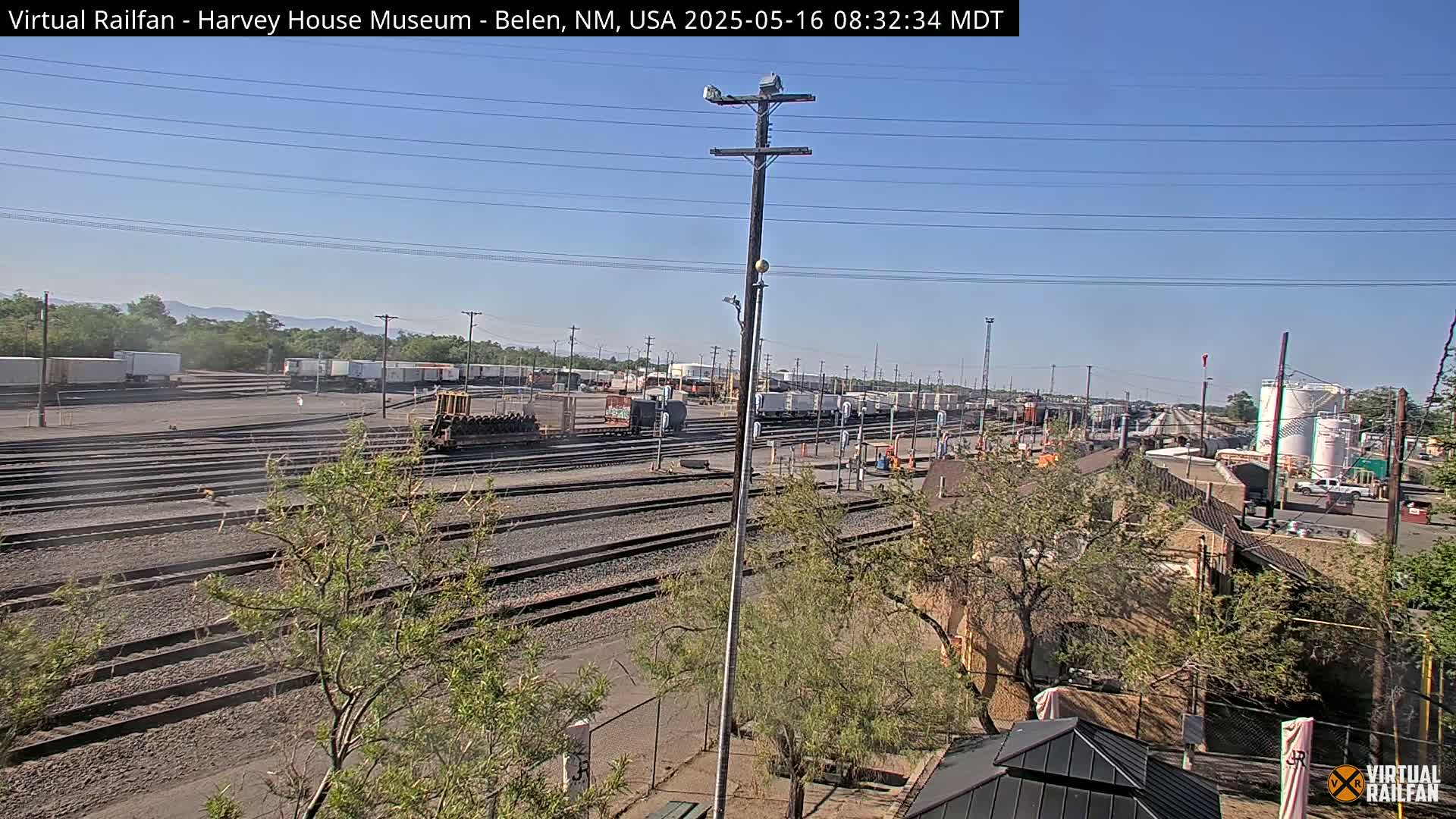 A sunny day reveals a sprawling rail yard with numerous train cars and tracks, some trees, and buildings in the distance.