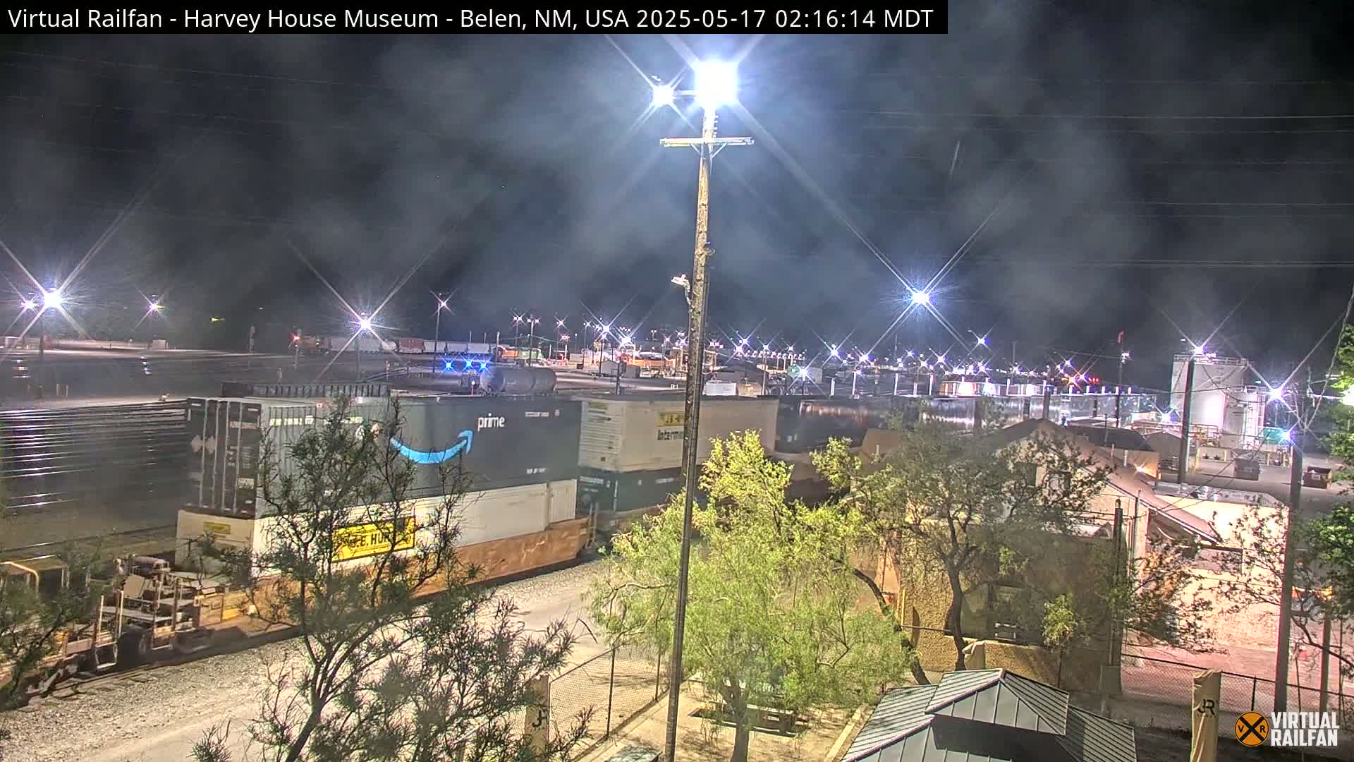 A nighttime view of a train carrying shipping containers, some marked with the Amazon Prime logo, illuminated by bright lights under a slightly hazy sky.