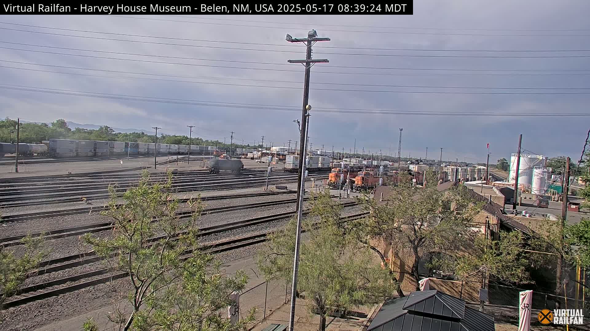 A mostly sunny day reveals a railway yard with multiple train cars and locomotives, surrounded by buildings and vegetation.