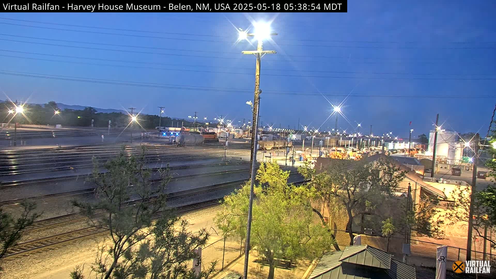 A twilight view of a rail yard with multiple train tracks, illuminated by artificial lights under a clear, dark blue sky.