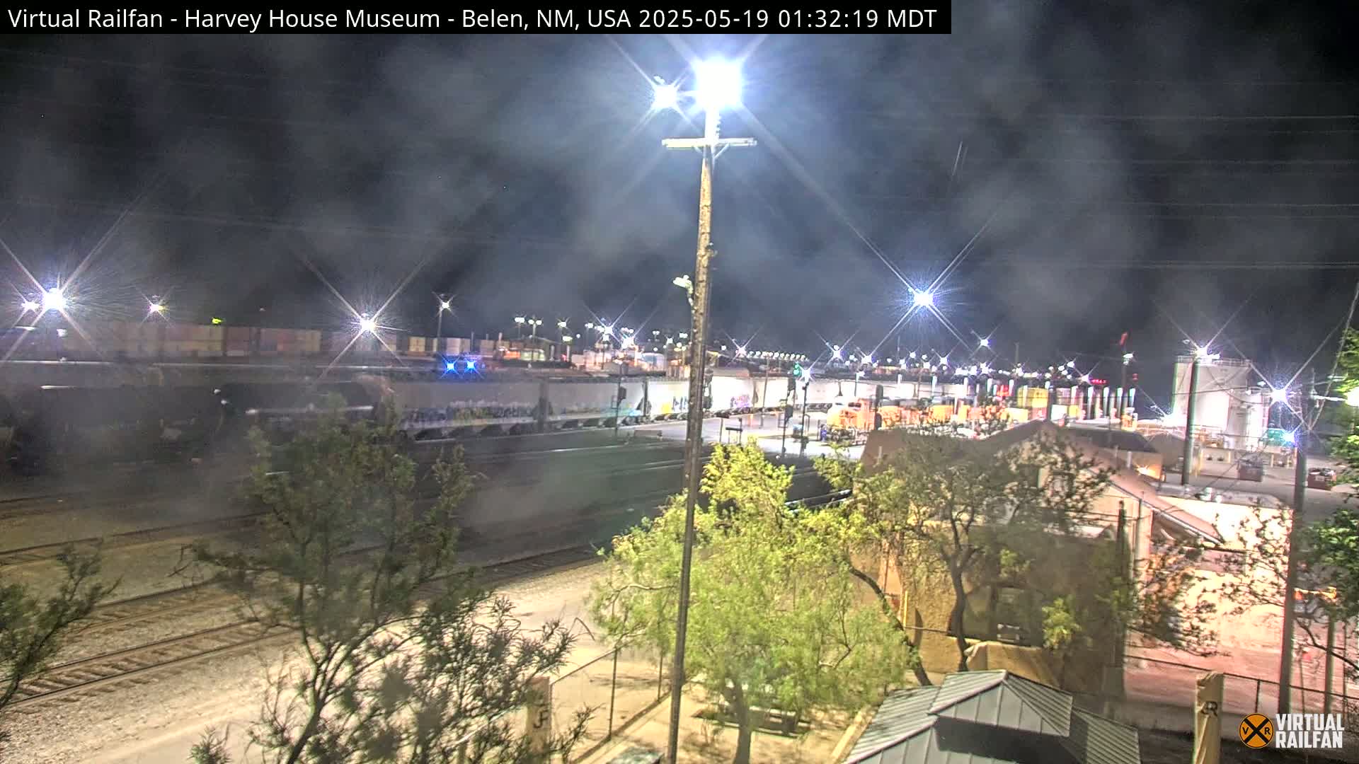 A nighttime view of a train yard with a long freight train, illuminated by bright lights, under a hazy sky.
