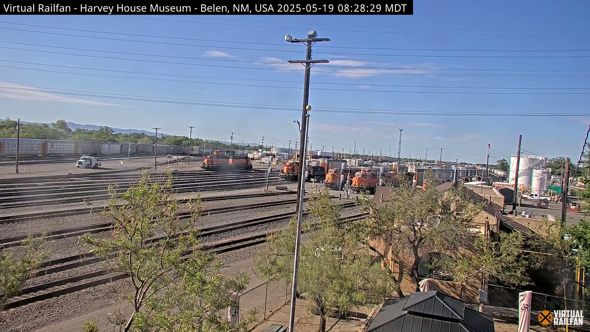A sunny day reveals a railroad yard with multiple orange locomotives and freight cars, alongside a white truck, and some low buildings partially obscured by trees.
