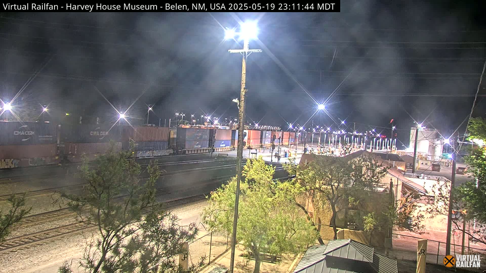 A nighttime view of a train carrying cargo containers, situated near buildings and trees under a hazy sky.