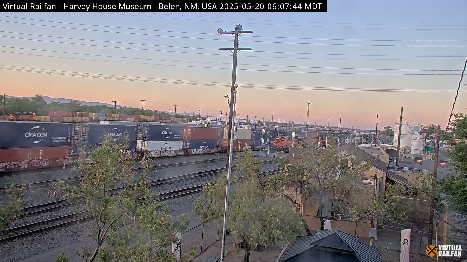 A freight train with multiple shipping containers is visible at dusk, with a pale pink sky and mountains in the background.
