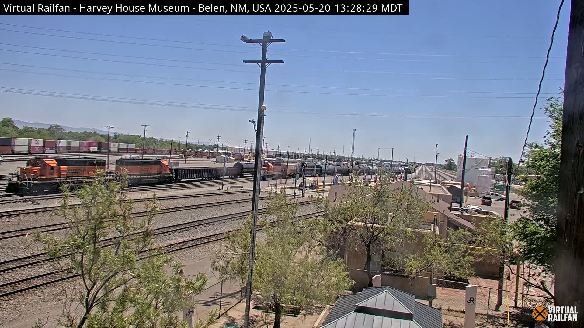 A sunny day at a rail yard shows orange locomotives and numerous freight cars parked along multiple sets of tracks, with some buildings and trees visible in the background.