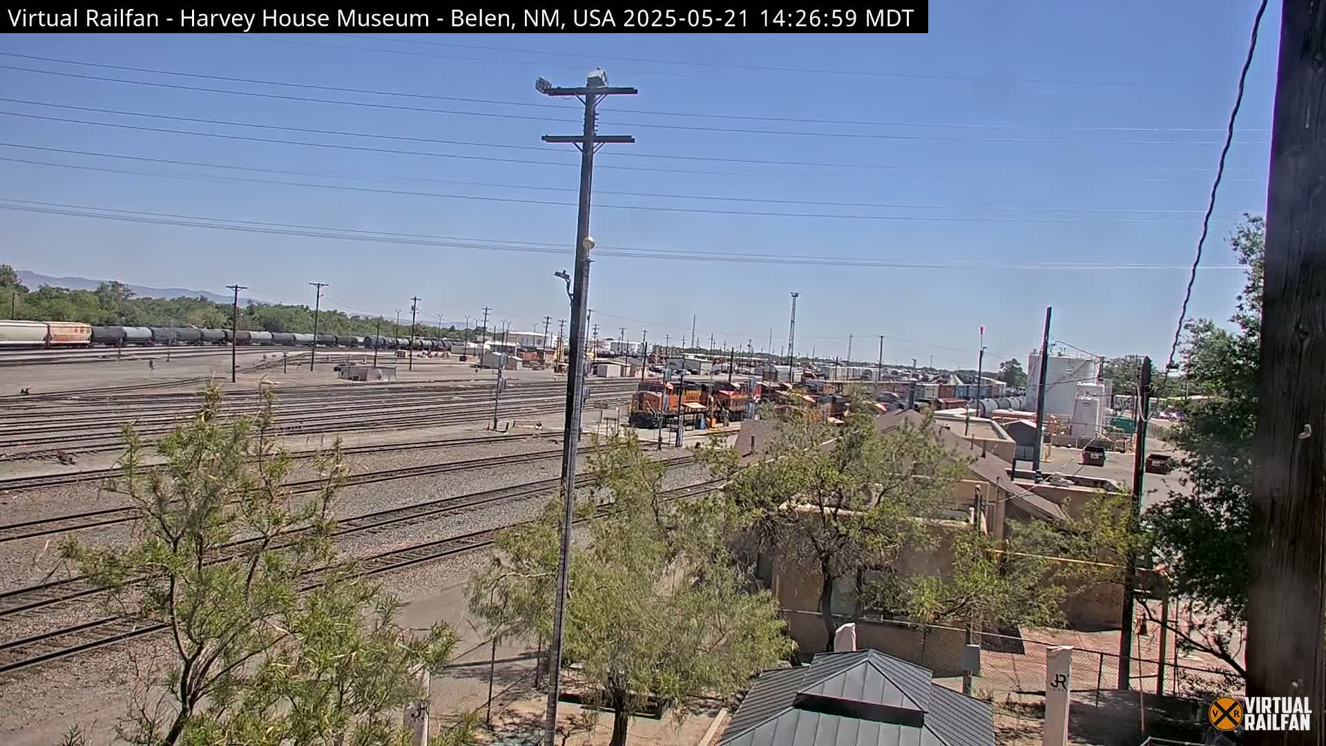 A sunny day reveals a sprawling rail yard with numerous train cars and tracks, surrounded by low desert vegetation and buildings.