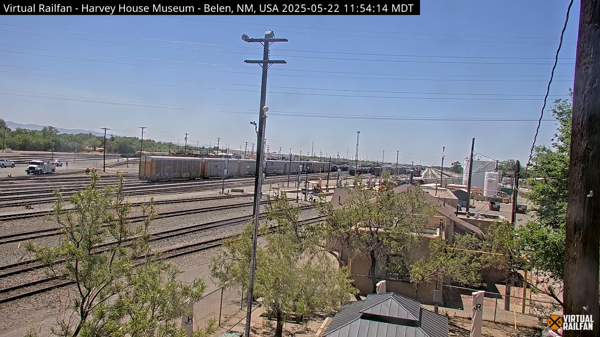 A sunny day reveals a wide expanse of railroad tracks with numerous freight cars, alongside some buildings and sparse vegetation.
