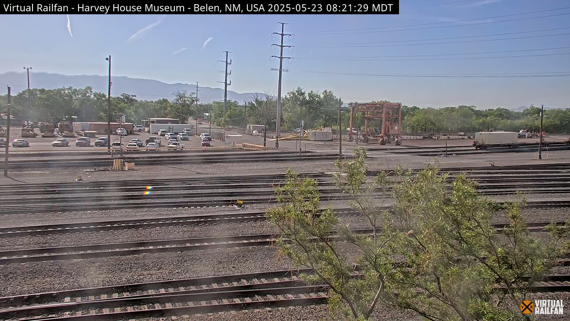 Under a clear sky, a train yard with multiple sets of tracks is visible, featuring a parking lot with several cars in the background and some trees and distant mountains.