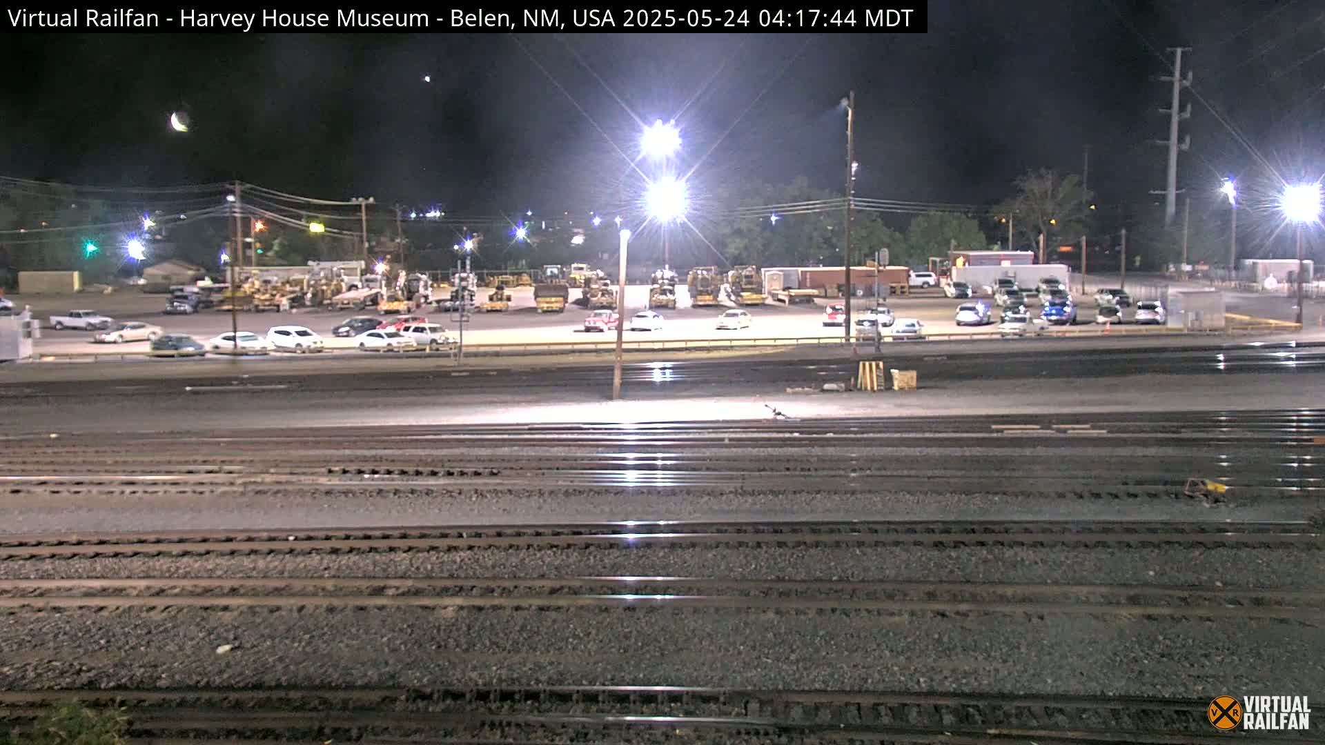 At night, under a crescent moon, several sets of train tracks run in the foreground, and beyond them are brightly lit parking lots filled with cars and construction equipment.