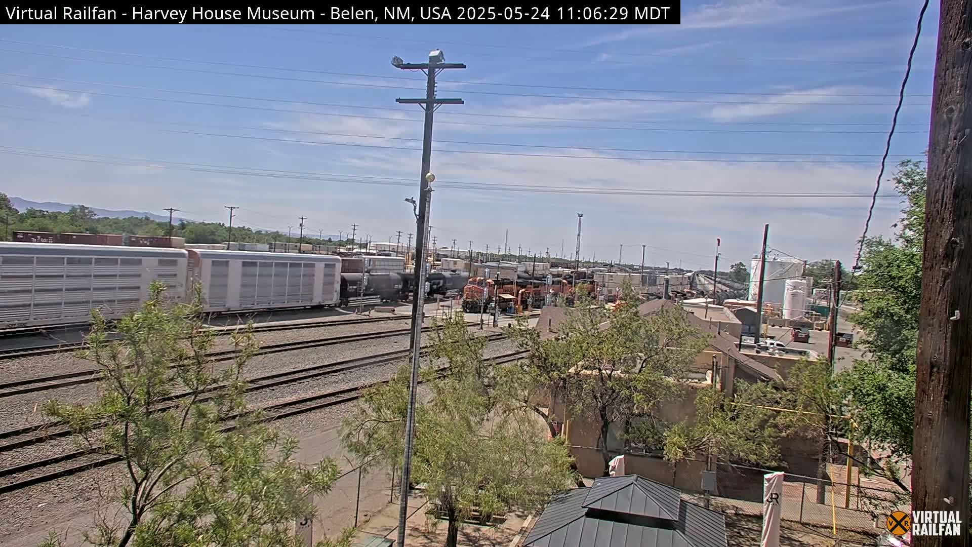 A mostly sunny day reveals a railroad yard with several train cars and locomotives, alongside some buildings and trees.