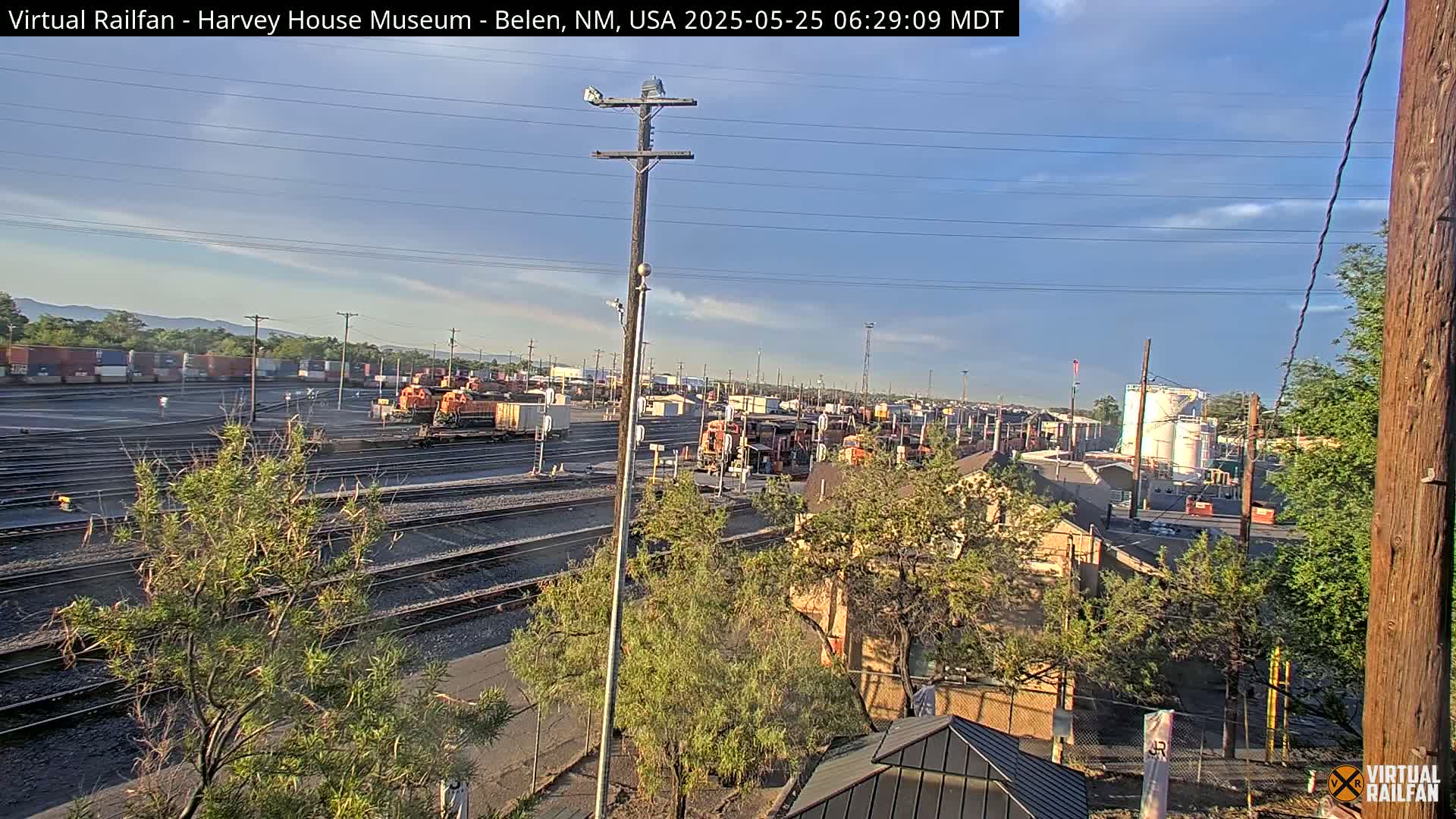 Under a mostly clear sky, a sprawling rail yard with multiple train cars and tracks is visible, surrounded by buildings and vegetation.