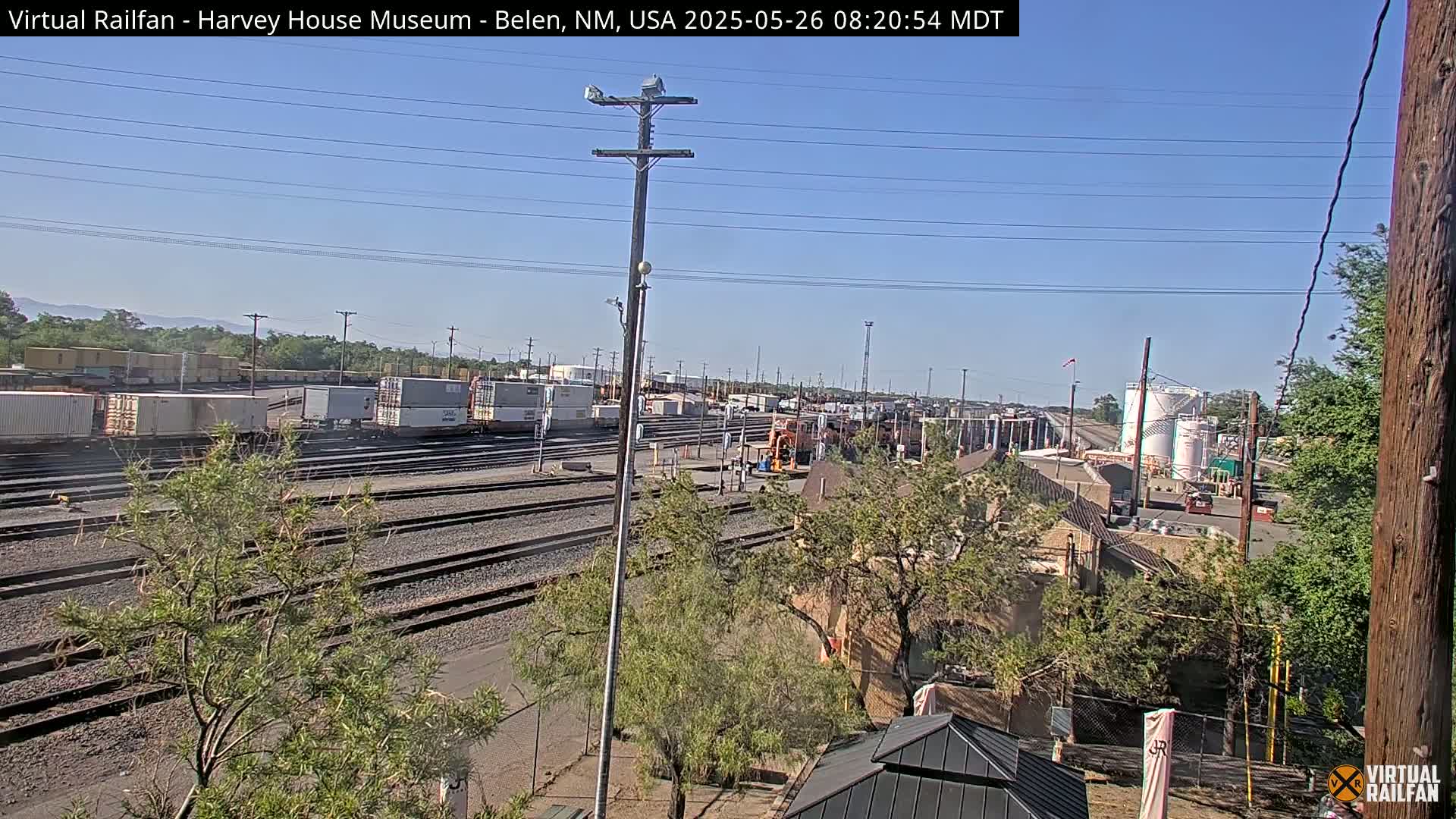 A sunny day reveals a train yard with numerous rail lines, freight cars, and a locomotive, situated amongst vegetation and buildings.