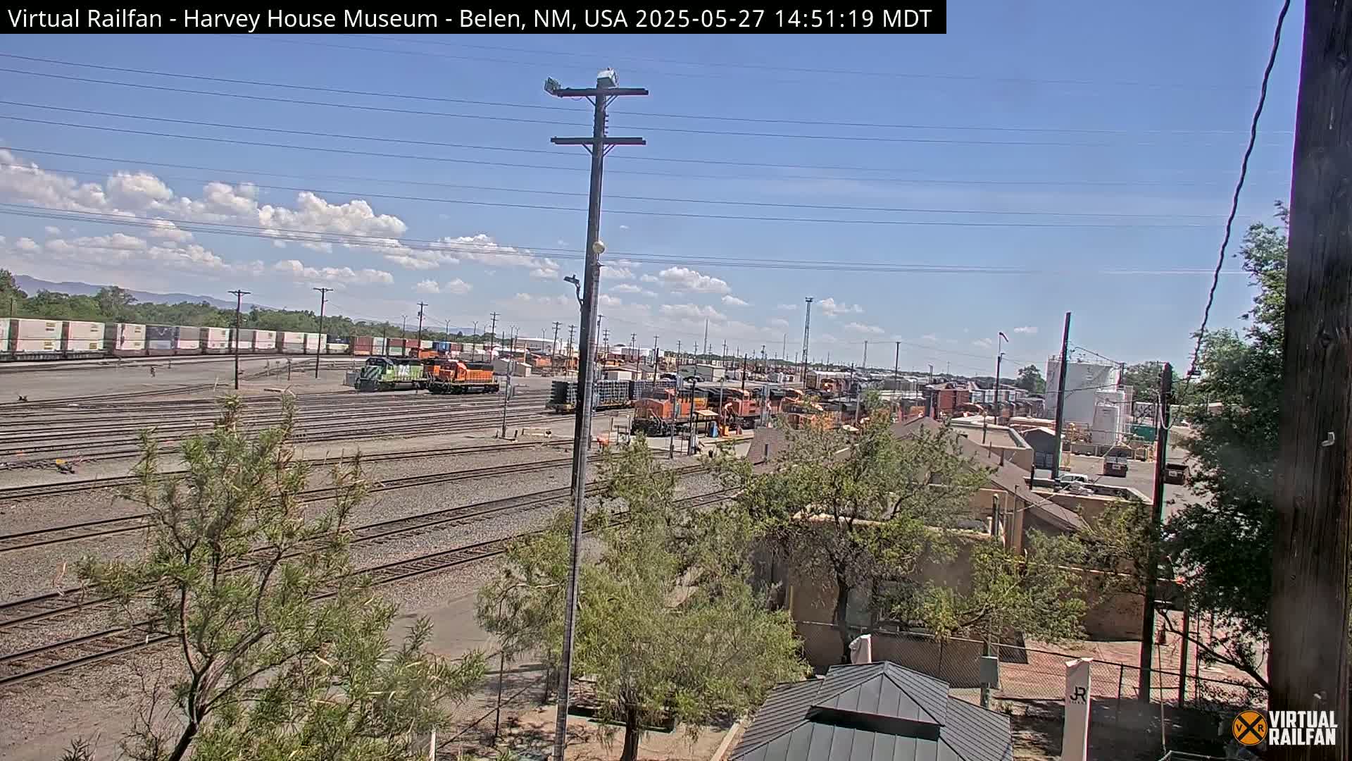 A sunny day reveals a large rail yard with several locomotives and freight cars, surrounded by trees and buildings.