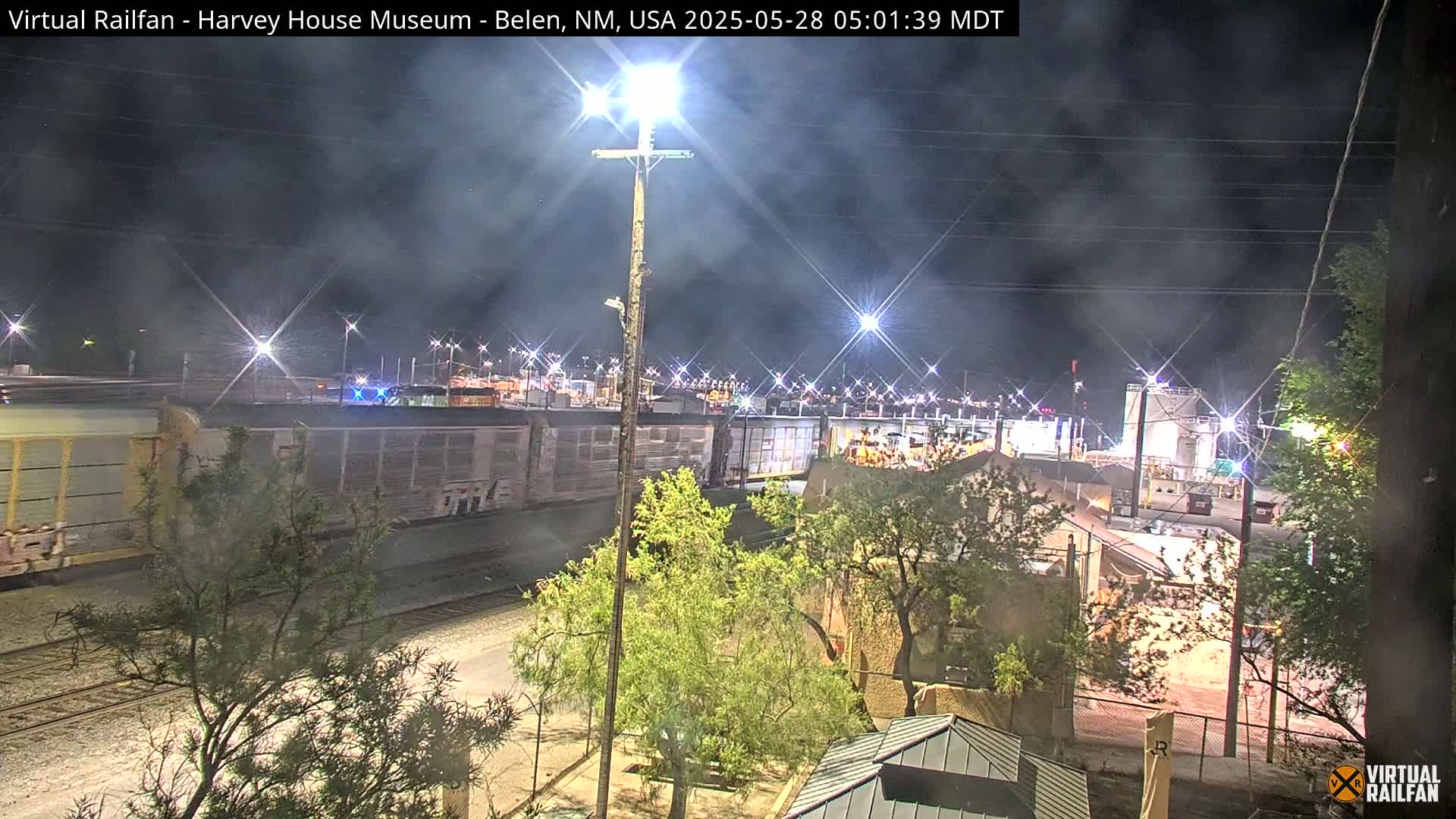 A nighttime view of a train passing by a rail yard, with hazy lighting conditions and trees in the foreground.