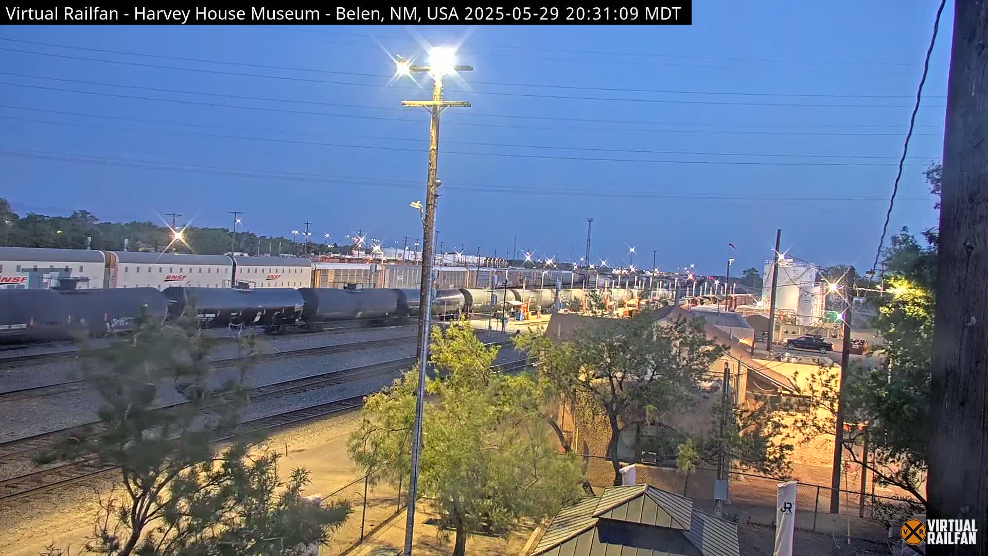 Under a twilight sky, a train of tanker cars sits on railroad tracks next to a rail yard and low buildings.