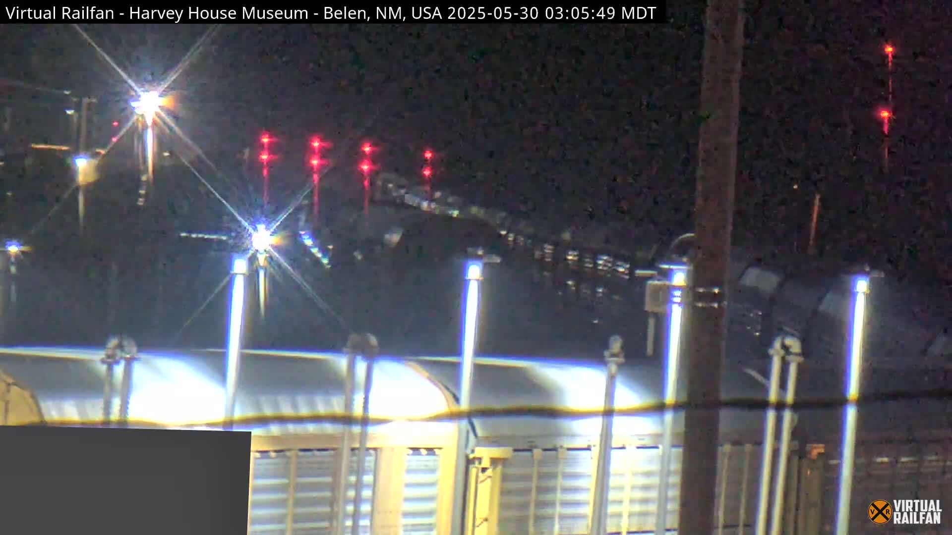 At night, a train passes by a brightly lit platform with several lights illuminating the scene.