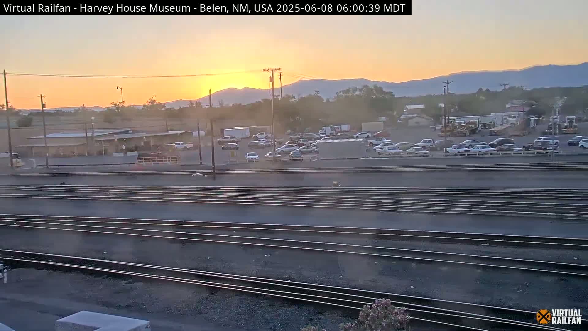 A sunset over a parking lot and railroad tracks with several cars and trucks visible in the lot.
