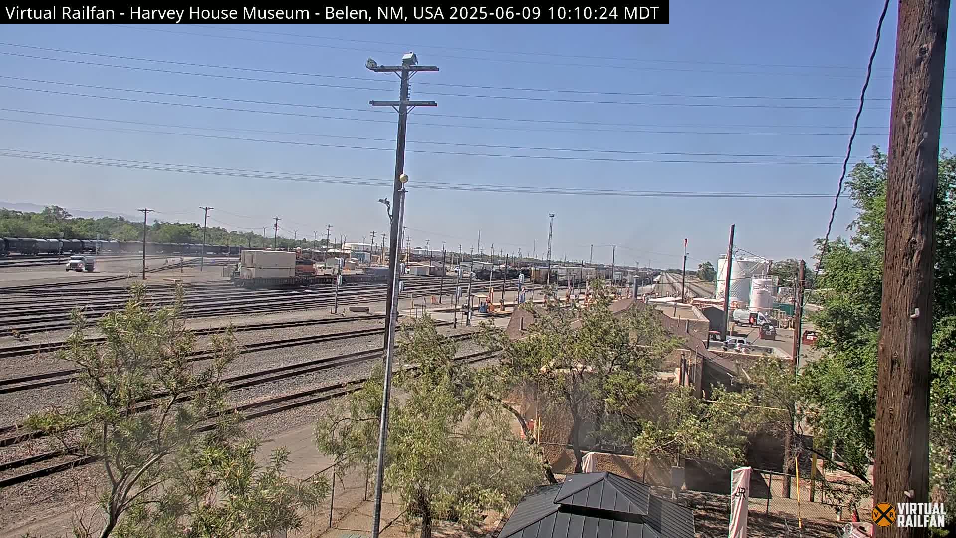 A sunny day reveals a sprawling railroad yard with numerous train cars, tracks, and surrounding vegetation.