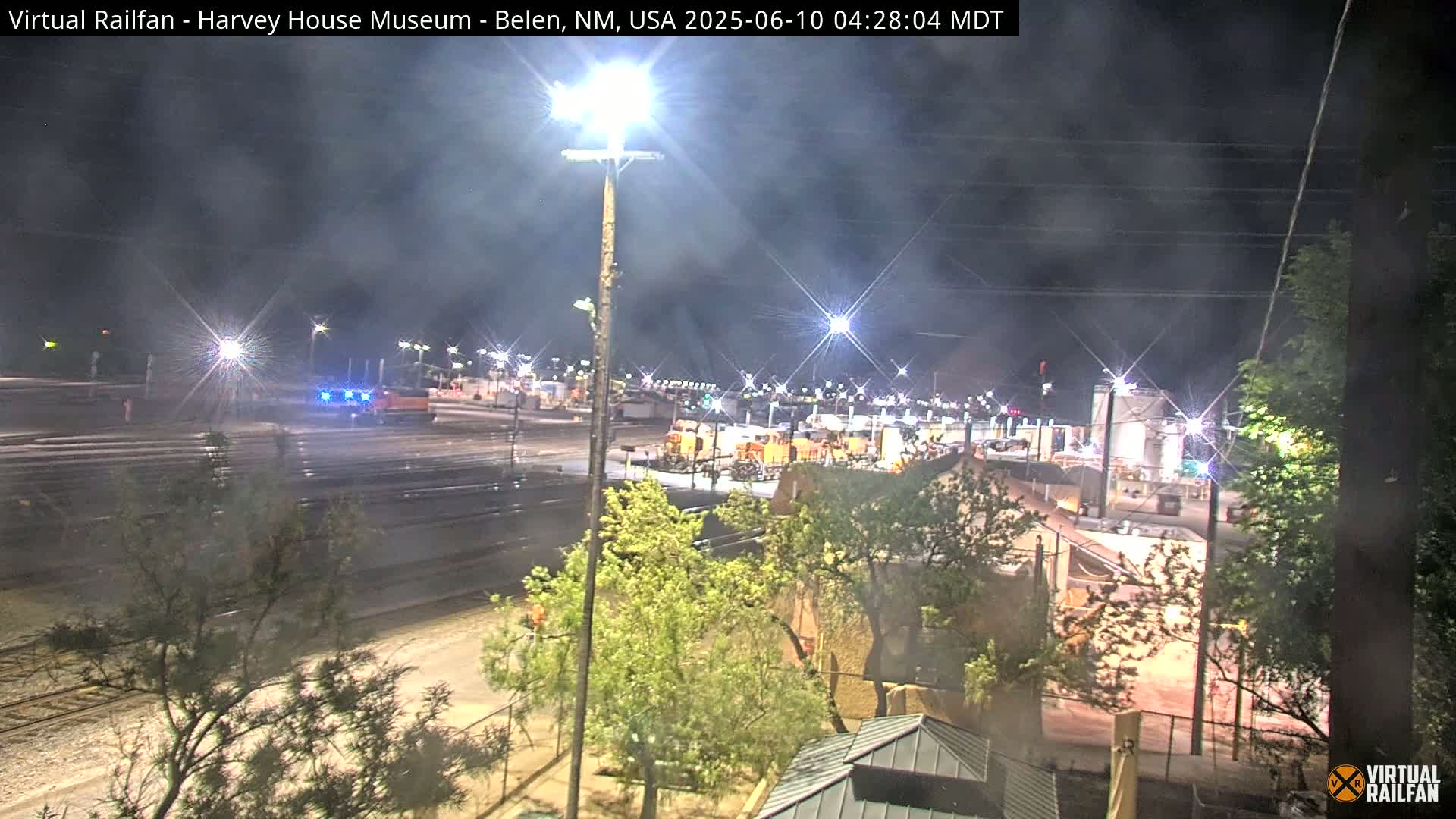 A nighttime view of a train yard with several locomotives and railcars illuminated under bright lights.