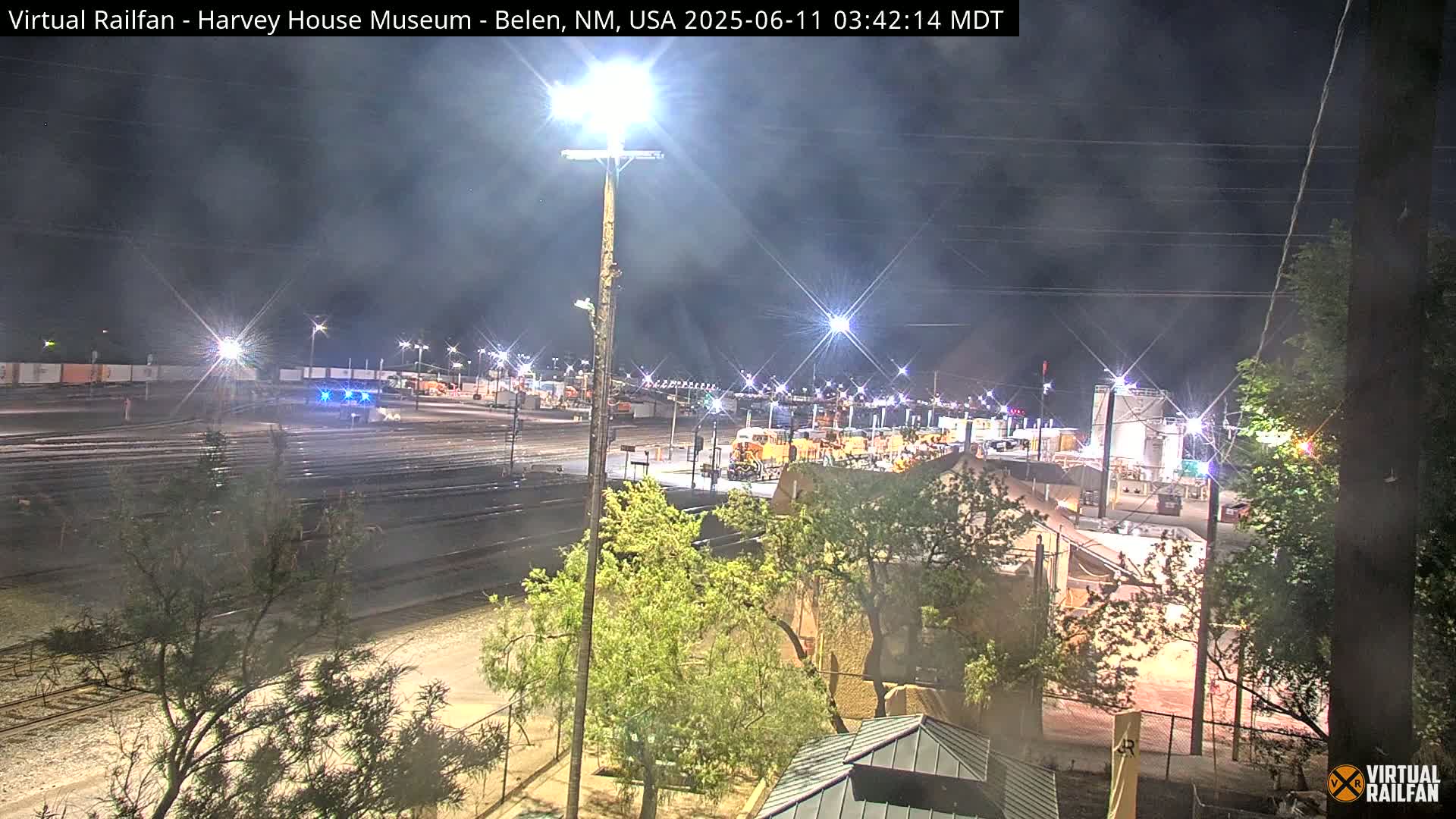 A nighttime view of a train yard with several locomotives and rail cars, under a partly cloudy sky.