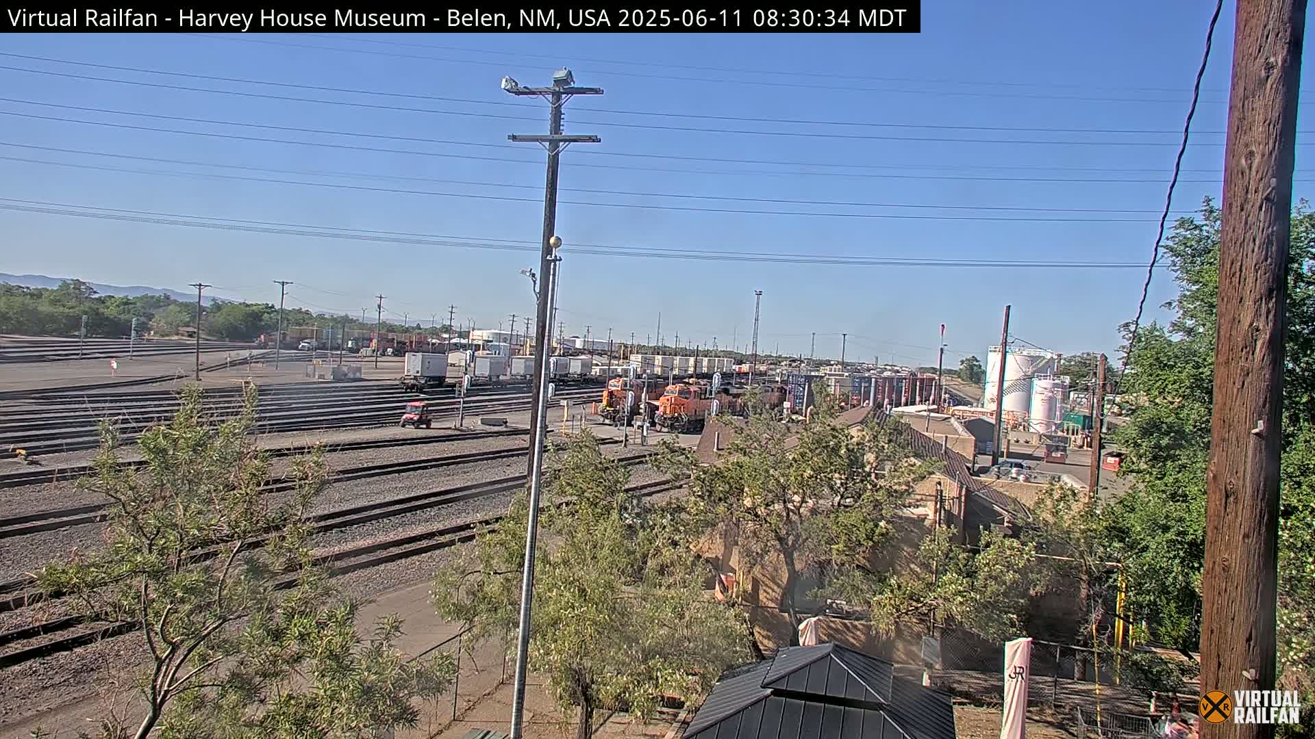 A sunny day reveals a sprawling railroad yard with multiple train cars and orange locomotives, situated amongst low-lying buildings and trees.